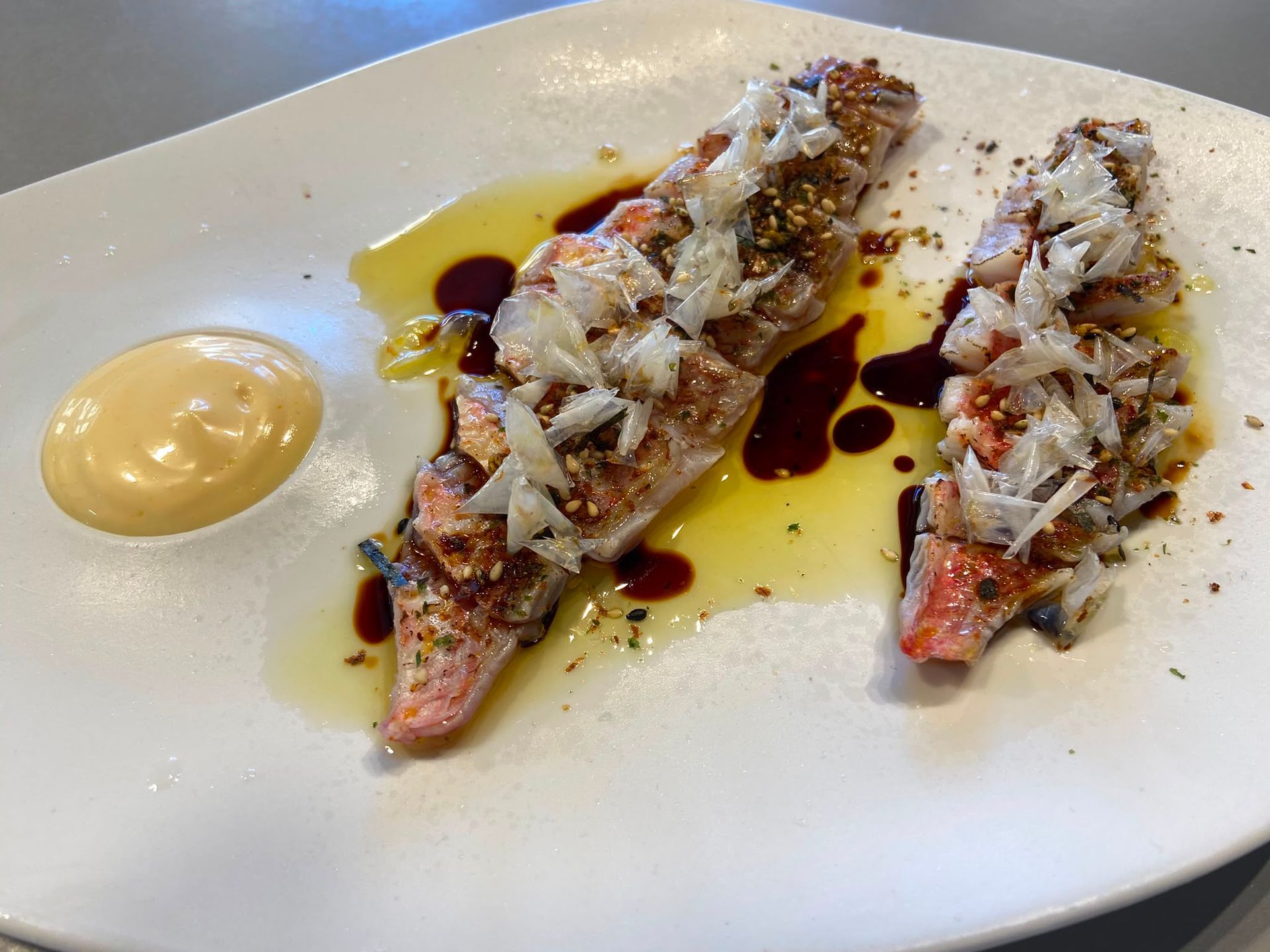 Aperitivo emplatado con dos piezas de marisco rellenas, bañadas en salsa, junto a una cucharada de alioli