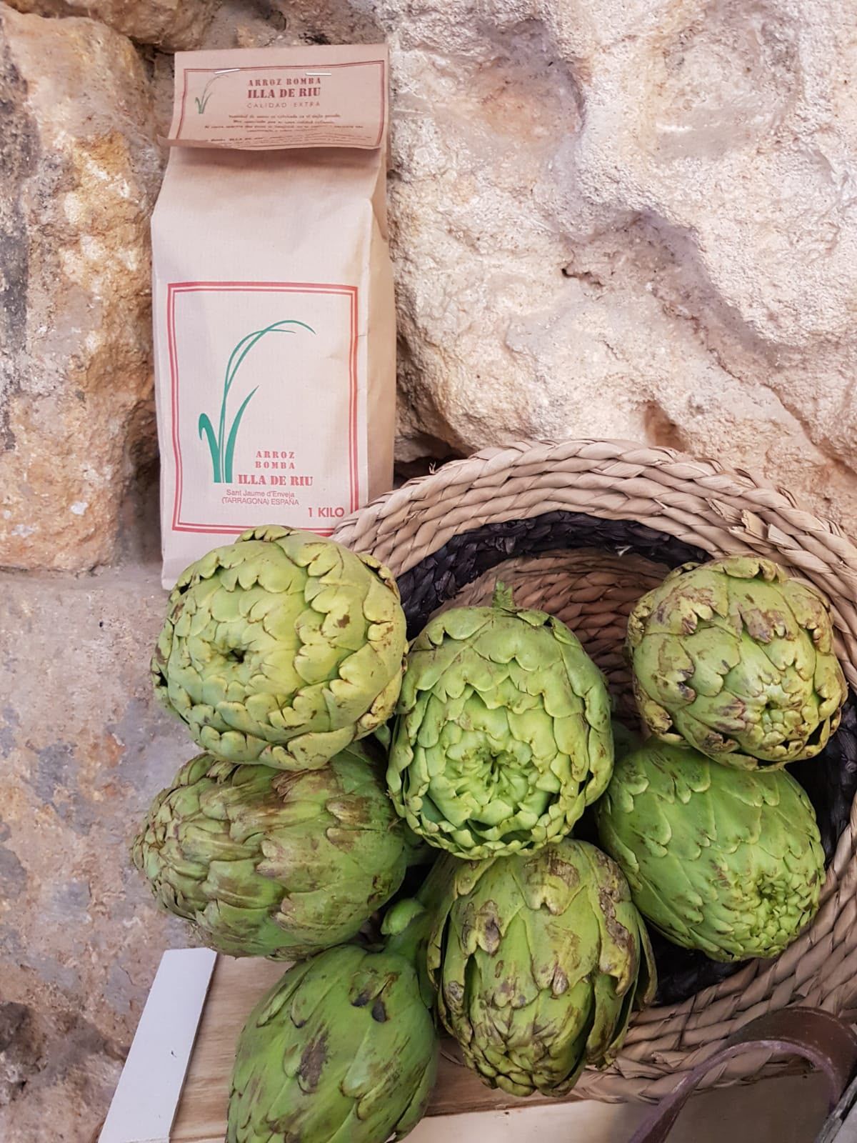 Cesta de alcachofas verdes junto a una bolsa de papel rosa sobre una superficie de piedra