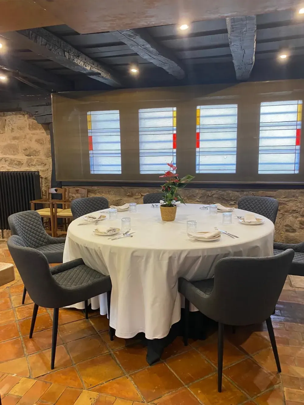 Mesa redonda puesta para una comida en un restaurante rústico, con sillas oscuras y persianas en las ventanas.