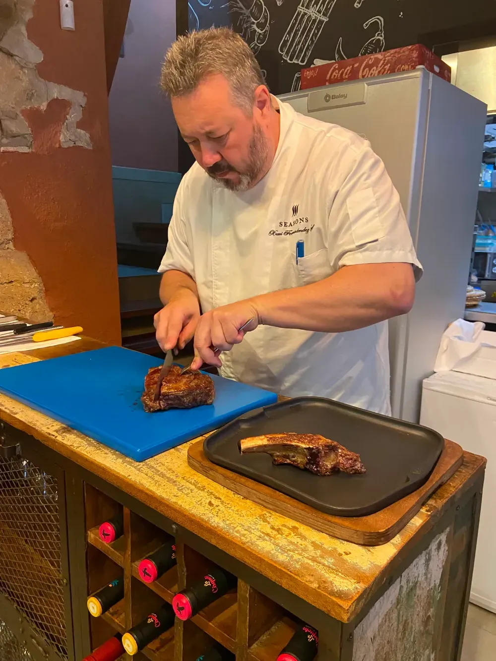 Un chef corta carne a la parrilla sobre una tabla de cortar azul, junto a una bandeja negra, en una cocina.