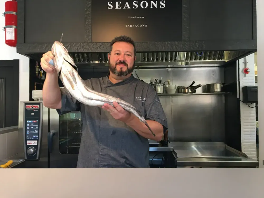 Un chef sostiene un pez grande en la cocina de un restaurante, debajo de un letrero que dice «SEASONS»
