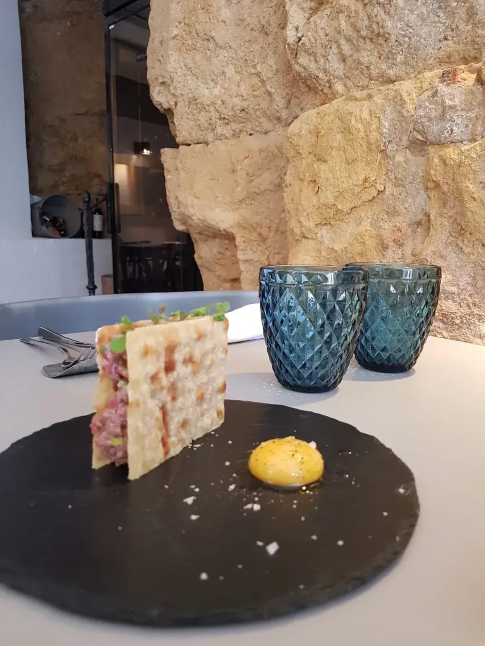 Postre gourmet emplatado en un plato negro, con copas azules, en un restaurante con paredes de piedra