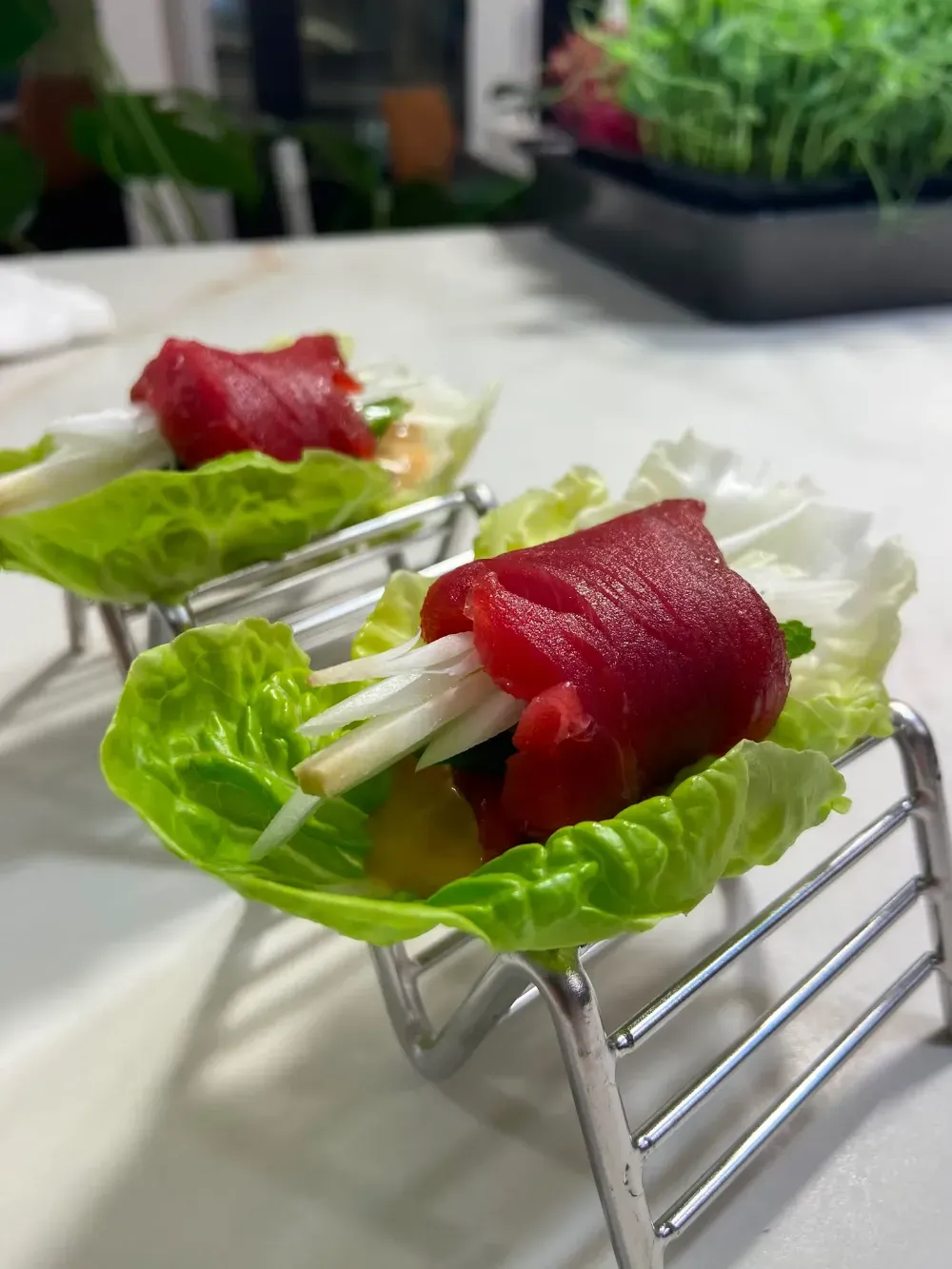 Hojas de lechuga rellenas de láminas de atún rojo sobre una rejilla metálica, con una cocina desenfocada al fondo