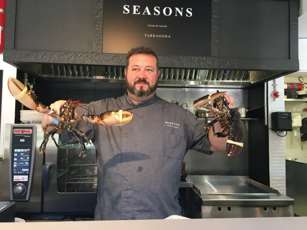 Chef con chaqueta gris sosteniendo dos langostas en la cocina de un restaurante