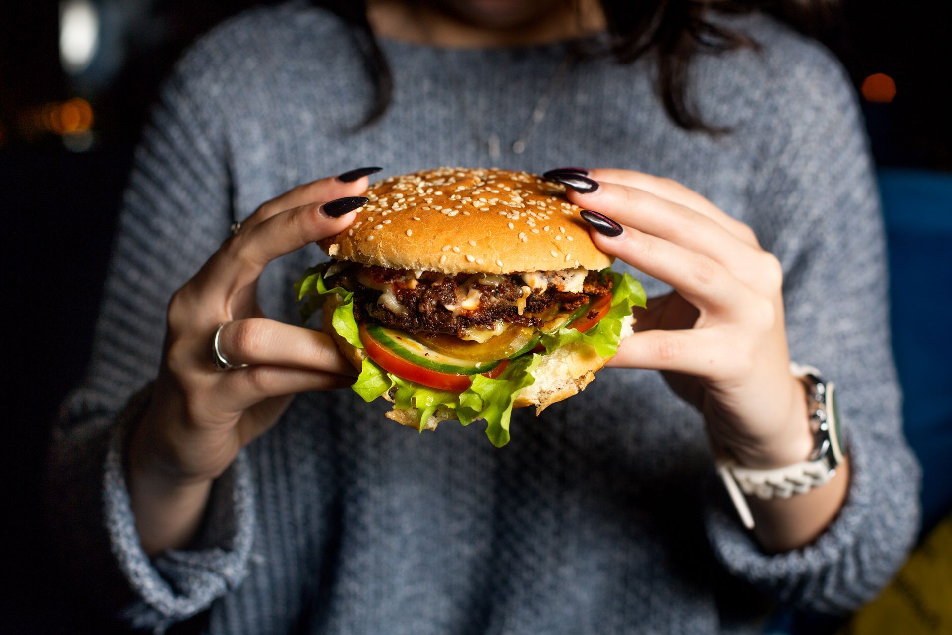 Persona sosteniendo una hamburguesa con pan con semillas de sésamo, lechuga, tomate y pepinillos.