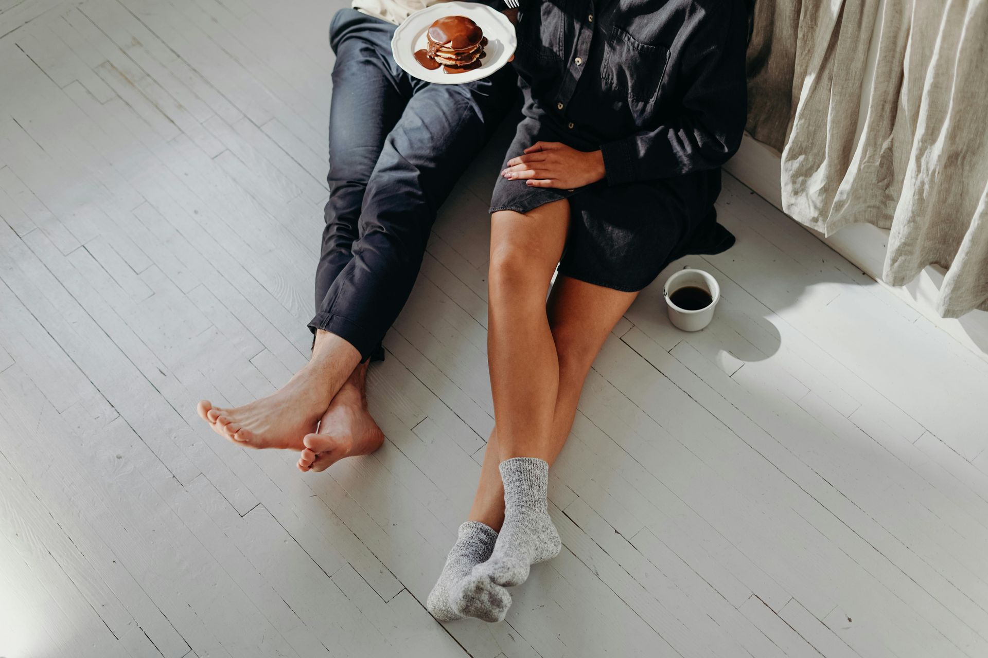 Pareja sentada en un suelo blanco, uno sosteniendo panqueques, el otro con café, entorno acogedor.