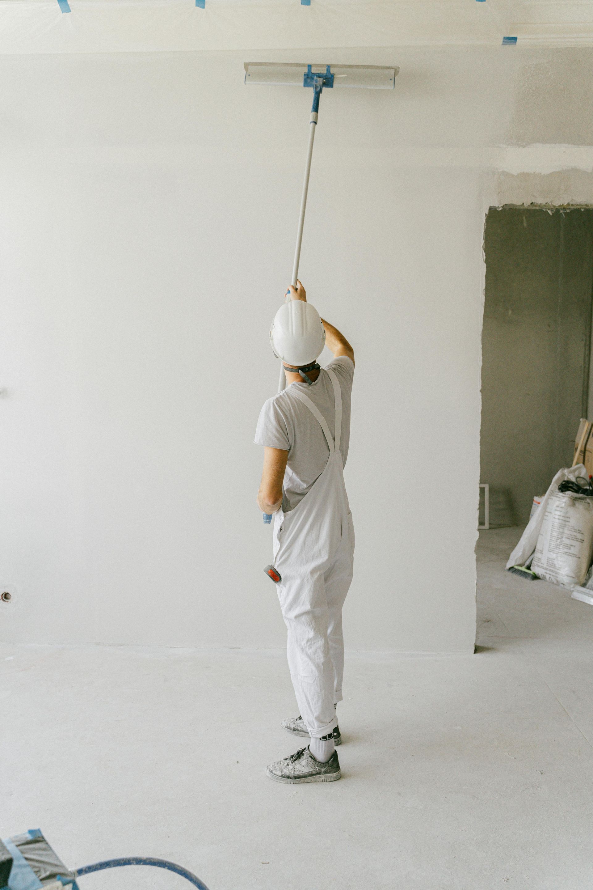 Persona con mono blanco y casco alisando un techo blanco con una herramienta larga en una habitación luminosa.