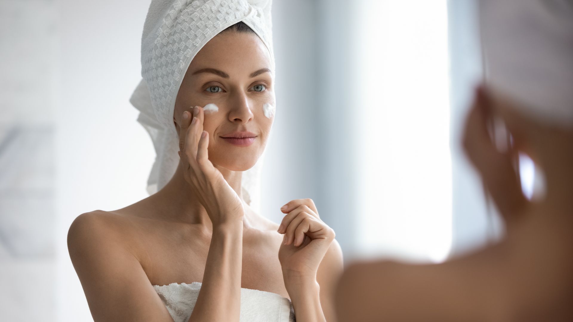 Une femme avec une serviette enroulée autour de sa tête applique de la crème sur son visage devant un miroir.