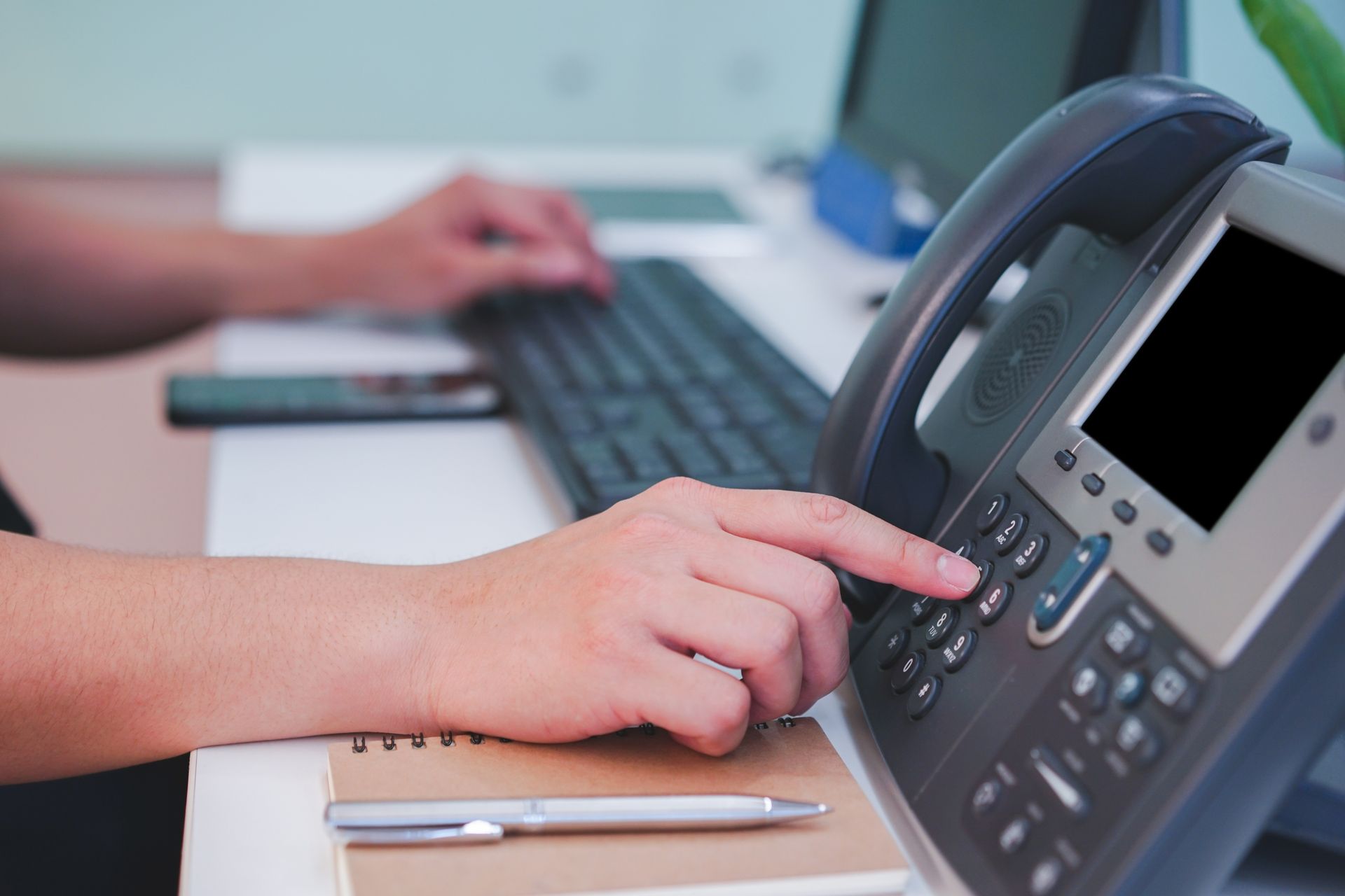 Une personne compose un numéro de téléphone sur un bureau. Un clavier et un ordinateur portable sont également présents, suggérant un environnement de bureau.