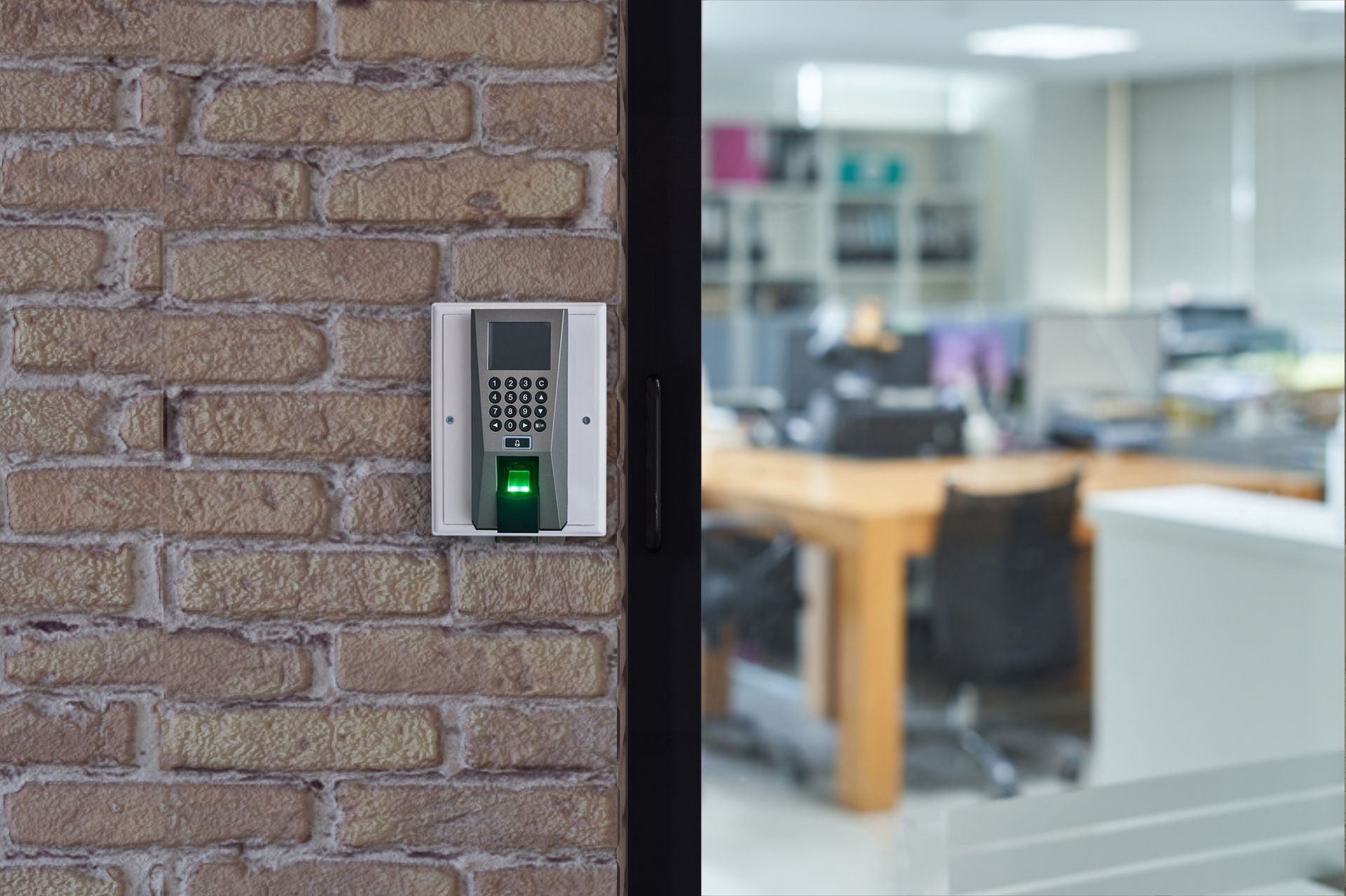 Scanner d'empreintes digitales sur un mur de briques, lumière verte allumée, bureau visible en arrière-plan.