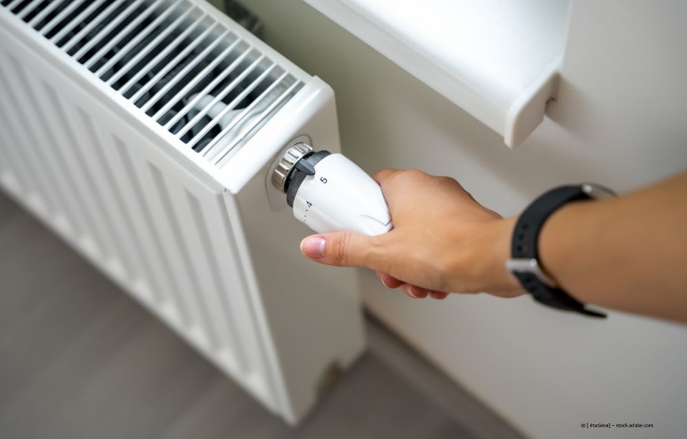 Eine Hand dreht den Thermostatknopf eines weißen Heizkörpers in der Nähe eines Fensters.