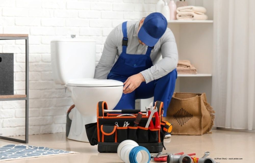 Ein Klempner in blauem Overall repariert eine Toilette in einem Badezimmer; sein Werkzeug liegt auf dem Boden.