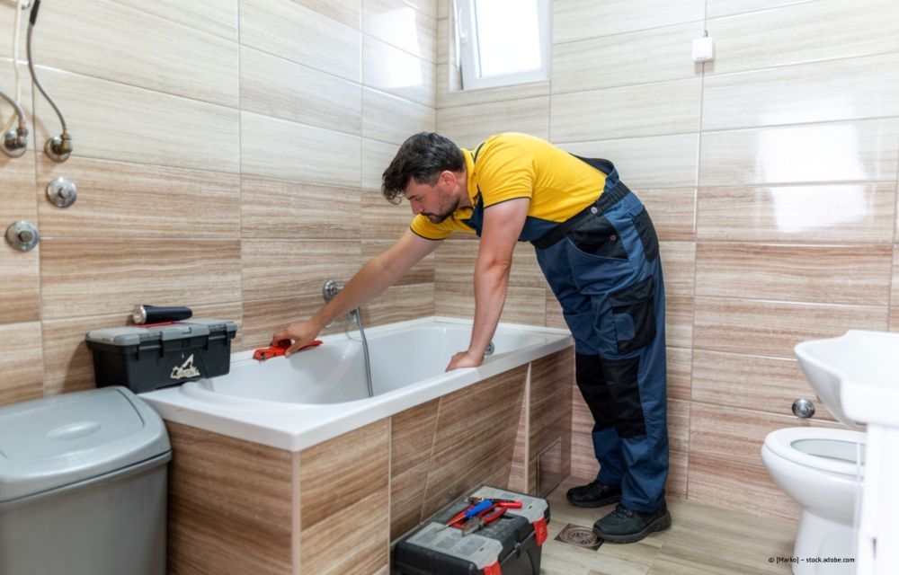 Ein Klempner repariert ein Waschbecken neben einer Toilette; er trägt ein gelbes Hemd und einen blauen Overall.
