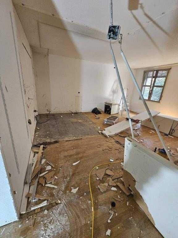 Zimmer im Umbau, Bodenbelag beschädigt, Bauschutt verstreut. Weisse Wände, Kleiderschrank, Fenster. Gelbes Verlängerungskabel.