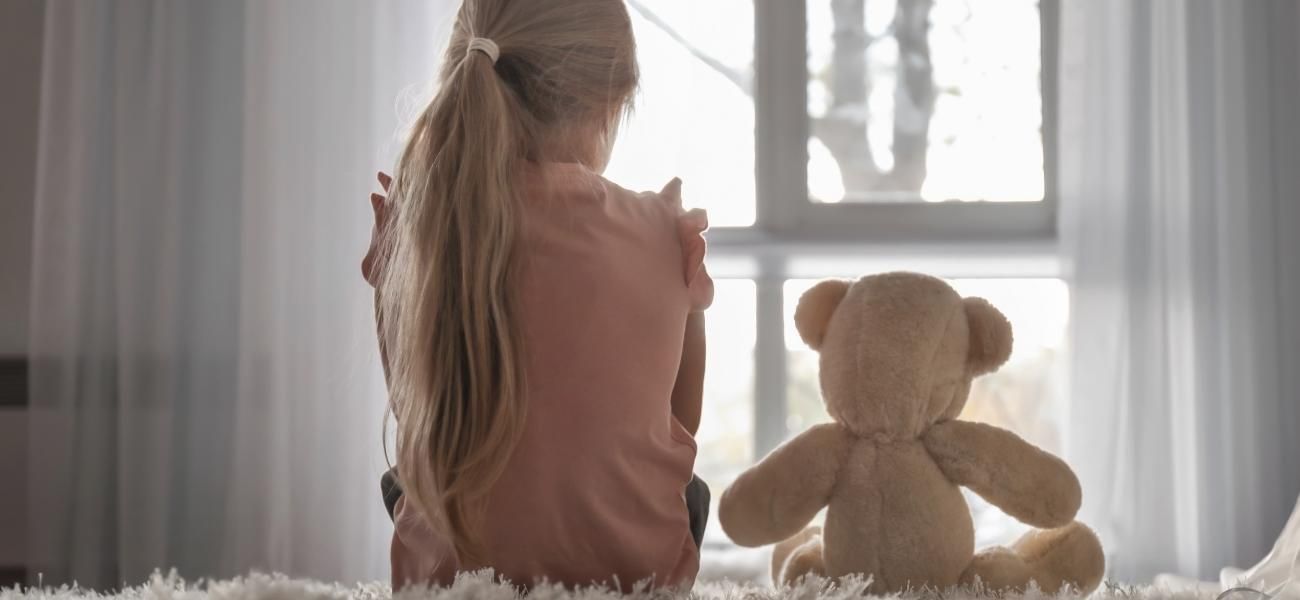 Una niña está sentada en el suelo junto a un osito de peluche.
