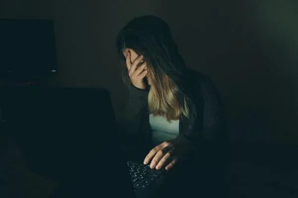 Una mujer se cubre la cara mientras usa una computadora portátil en la oscuridad.