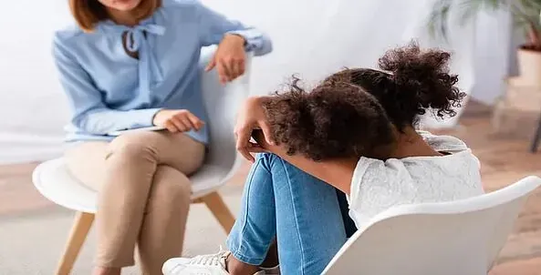 Una mujer está sentada en una silla hablando con una niña que está sentada en una silla con la cabeza gacha.