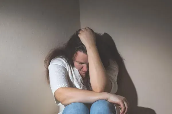 Una mujer está sentada en un rincón de una habitación con la cabeza entre las manos.