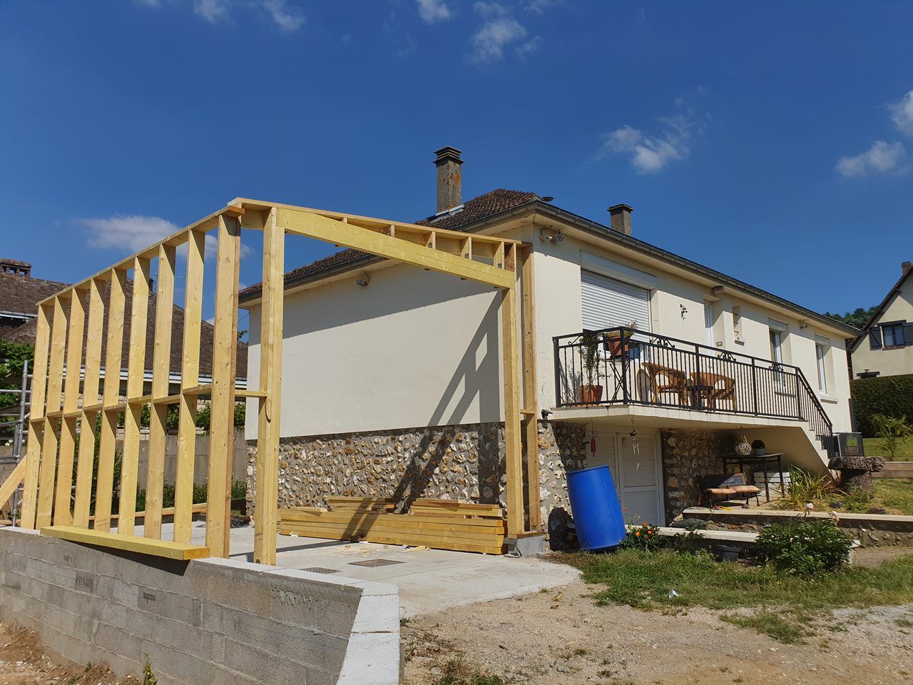 Charpente de porche en bois partiellement construite à côté d'une maison blanche sous un ciel bleu