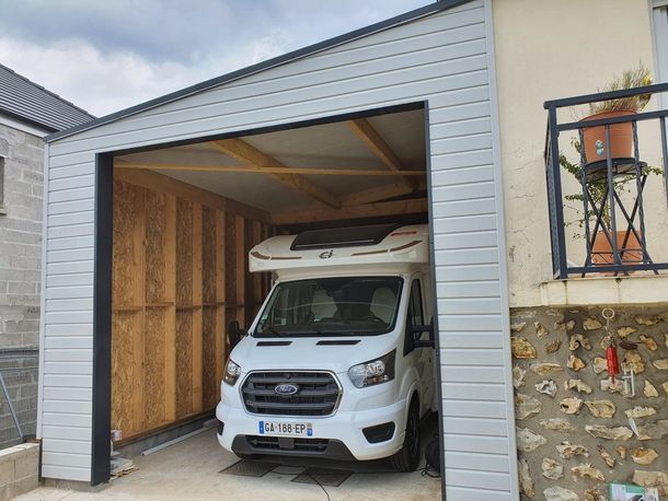 Une camionnette blanche garée dans un petit garage, porte ouverte, à côté d'une maison en pierre.