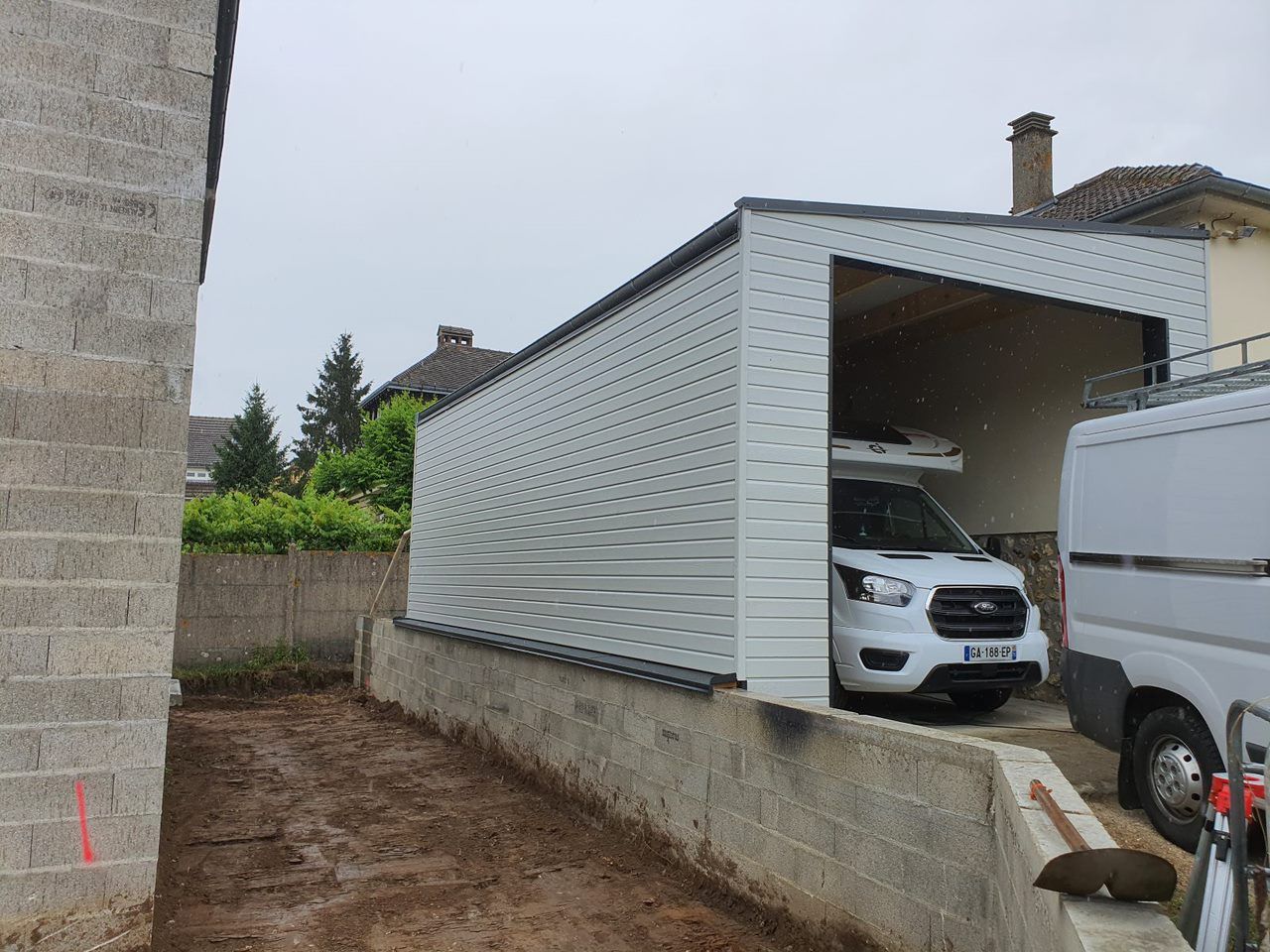 Une camionnette blanche est garée sous un étroit abri de voiture, à côté d'un mur de pierre et de l'extérieur d'une maison.