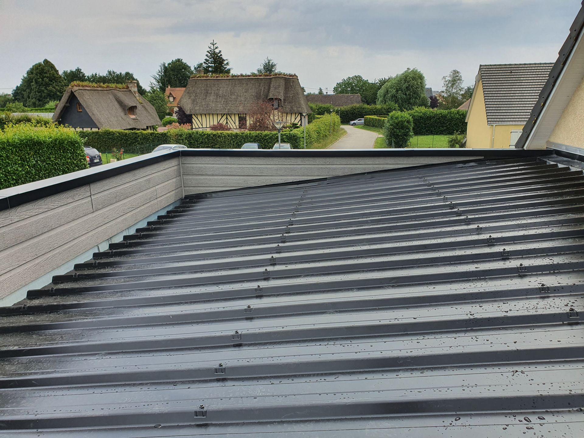Terrasse sur le toit avec toit en tôle ondulée grise, haies et maisons au loin sous un ciel nuageux