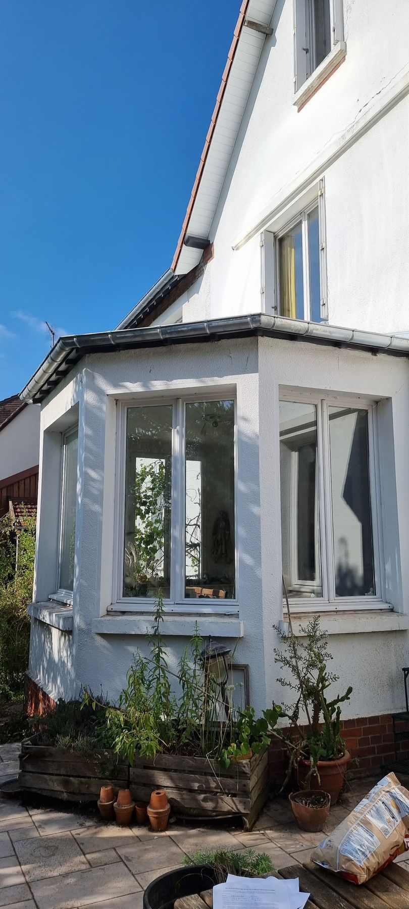 Extérieur d'une maison blanche avec une baie vitrée, des fondations en briques, des plantes en pot et un ciel bleu.