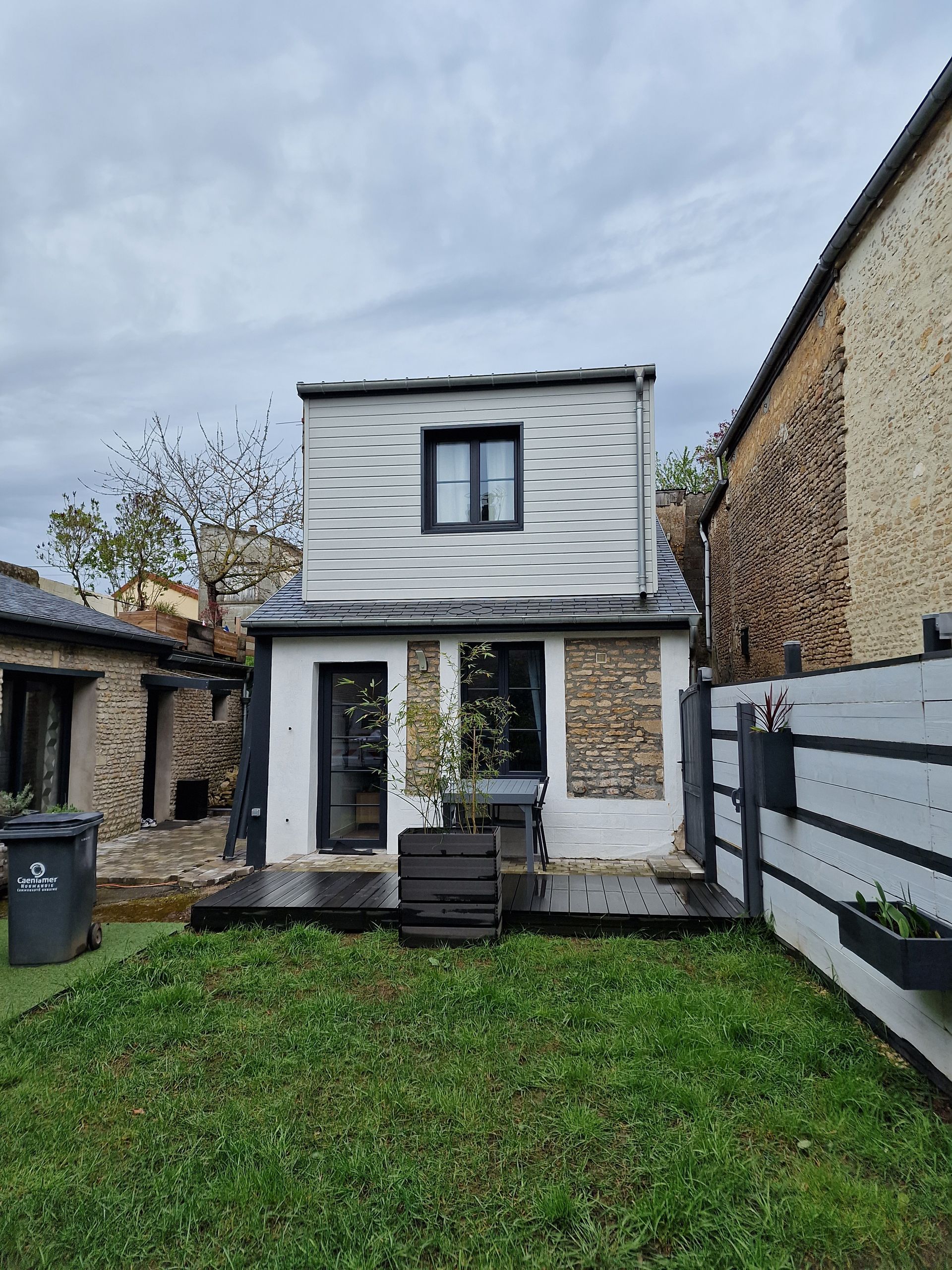 Petite maison blanche à deux étages avec une cour gazonnée, des murs en pierre et un ciel nuageux.
