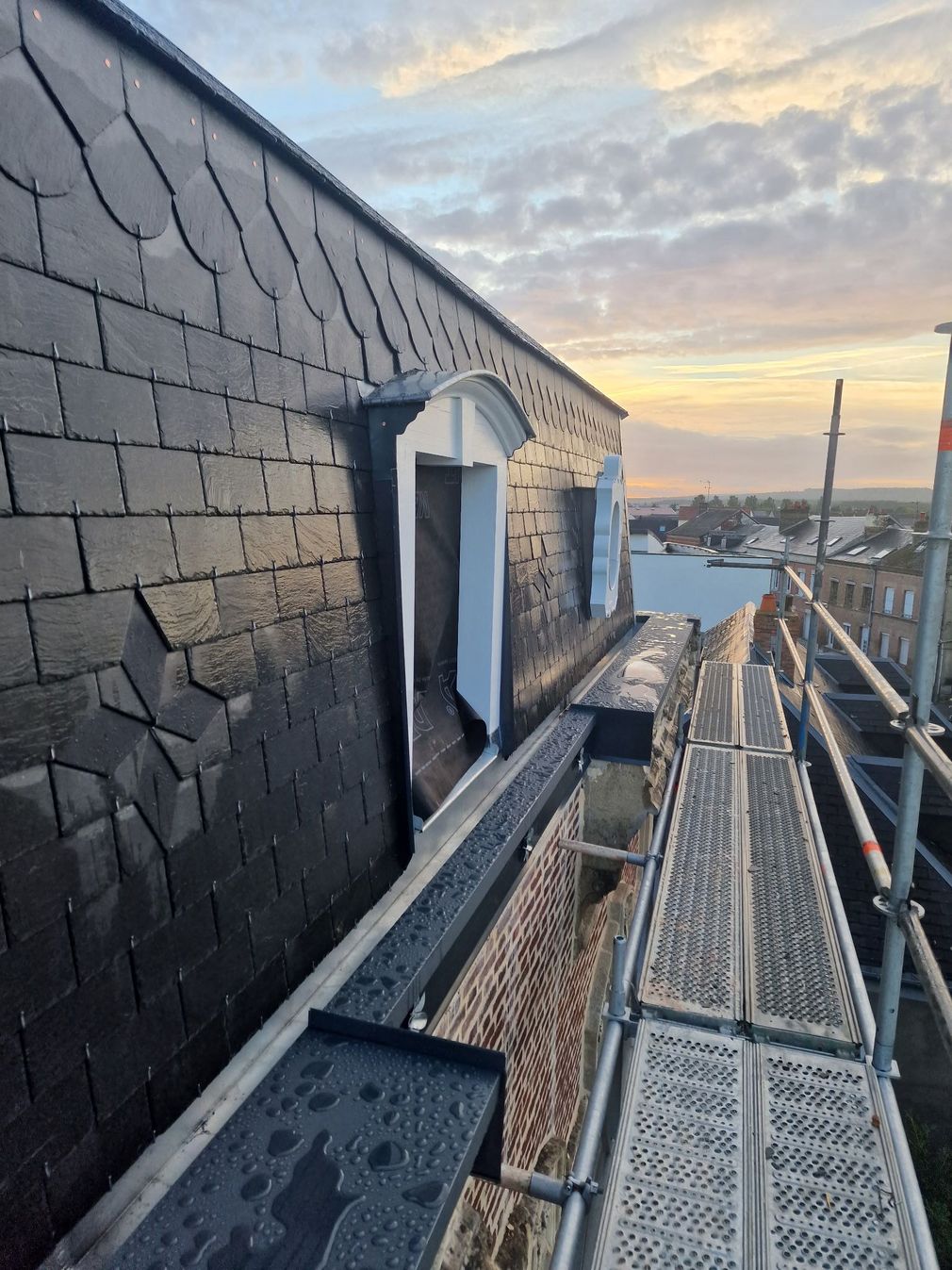Passerelle sur le toit avec une grille métallique longeant un mur d'ardoise au coucher du soleil, donnant sur l'eau et une tour au loin.