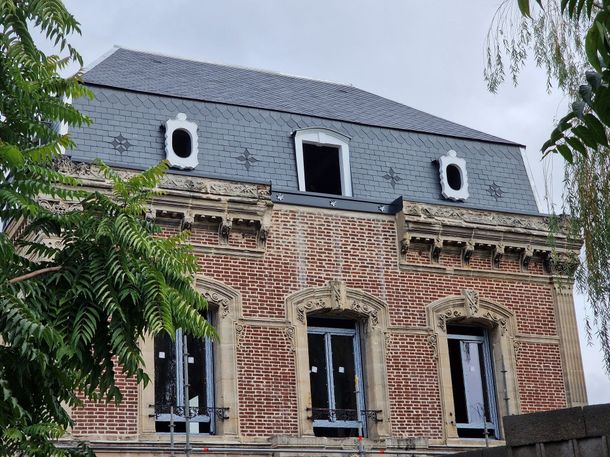 Façade d'une vieille maison en briques avec un toit d'ardoise sombre et plusieurs fenêtres, partiellement encadrées par des branches d'arbres