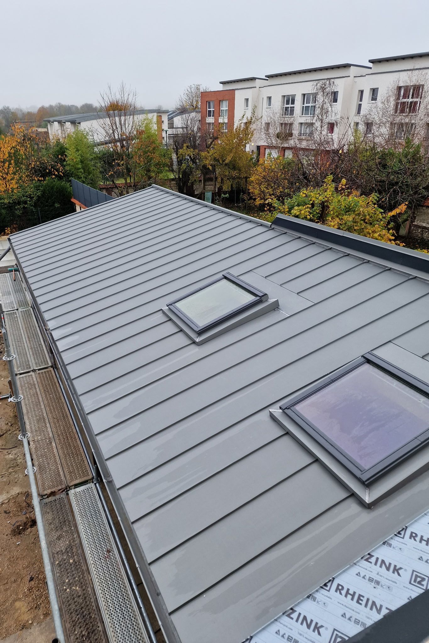 Toiture métallique grise avec deux puits de lumière, vue sur les arbres et les immeubles d'appartements sous un ciel couvert