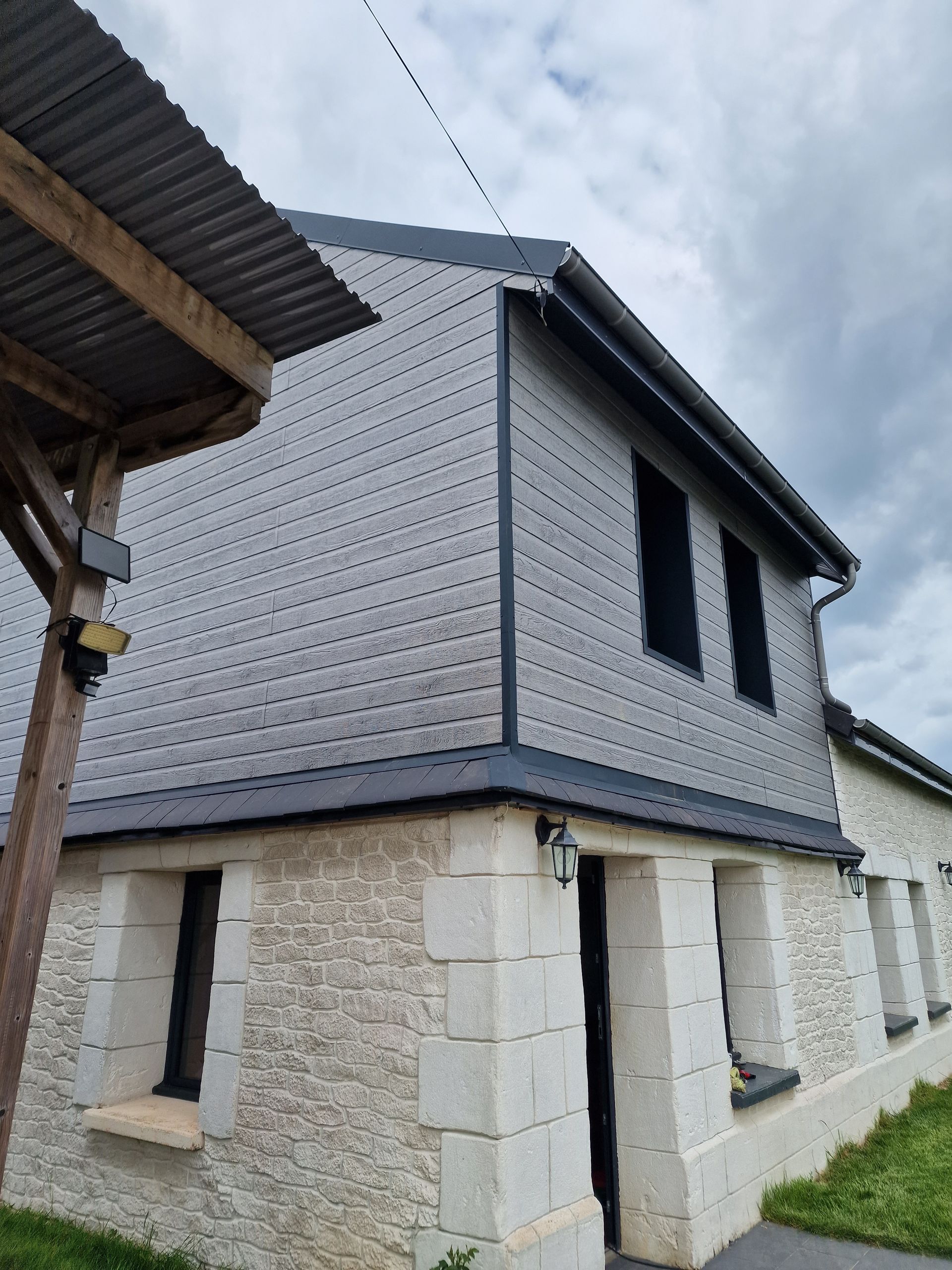 Maison en pierre de deux étages avec un bardage supérieur gris, des volets noirs et un toit en pente.