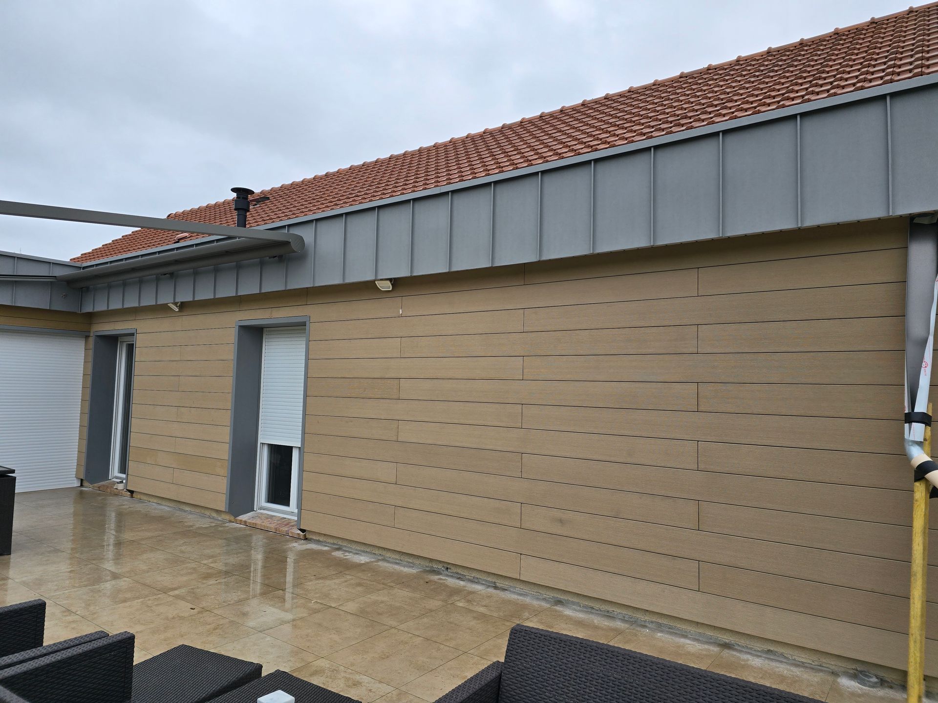 Maison en briques avec toiture grise et terrasse ombragée devant des portes et fenêtres blanches.