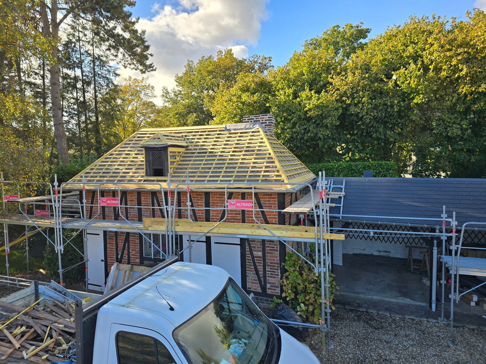 Maison en construction avec échafaudages, charpente de toit et une camionnette blanche devant.