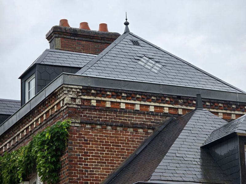Bâtiment en briques avec un toit en ardoise grise, des lucarnes et du lierre sous un ciel couvert