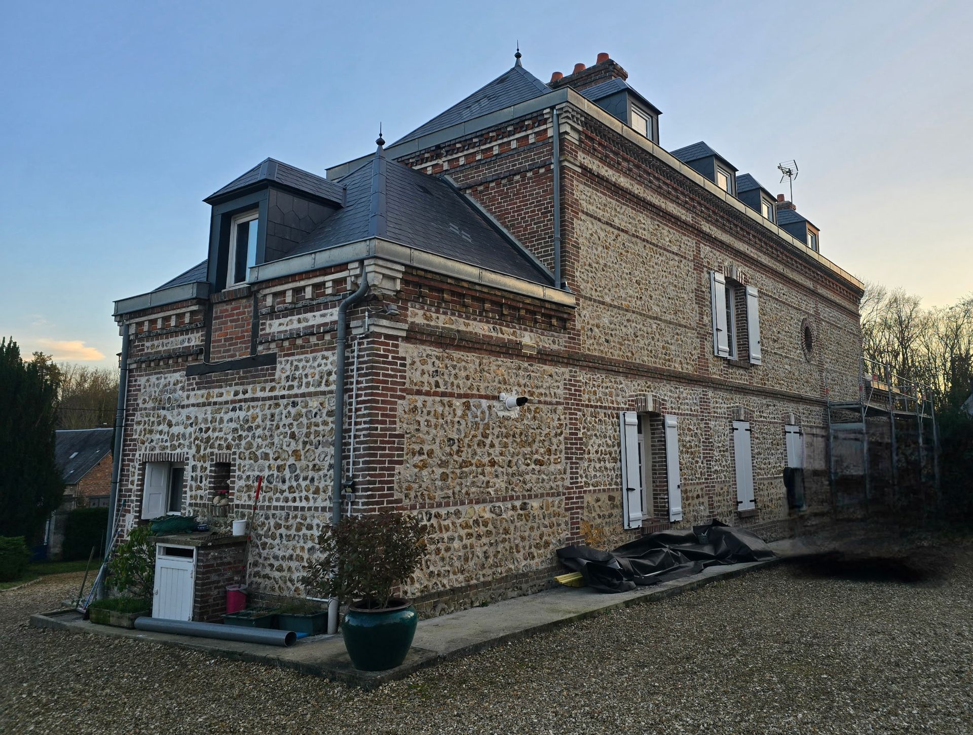 Maison en pierre avec un toit en ardoise et des volets blancs, au bord d'une allée de gravier au crépuscule