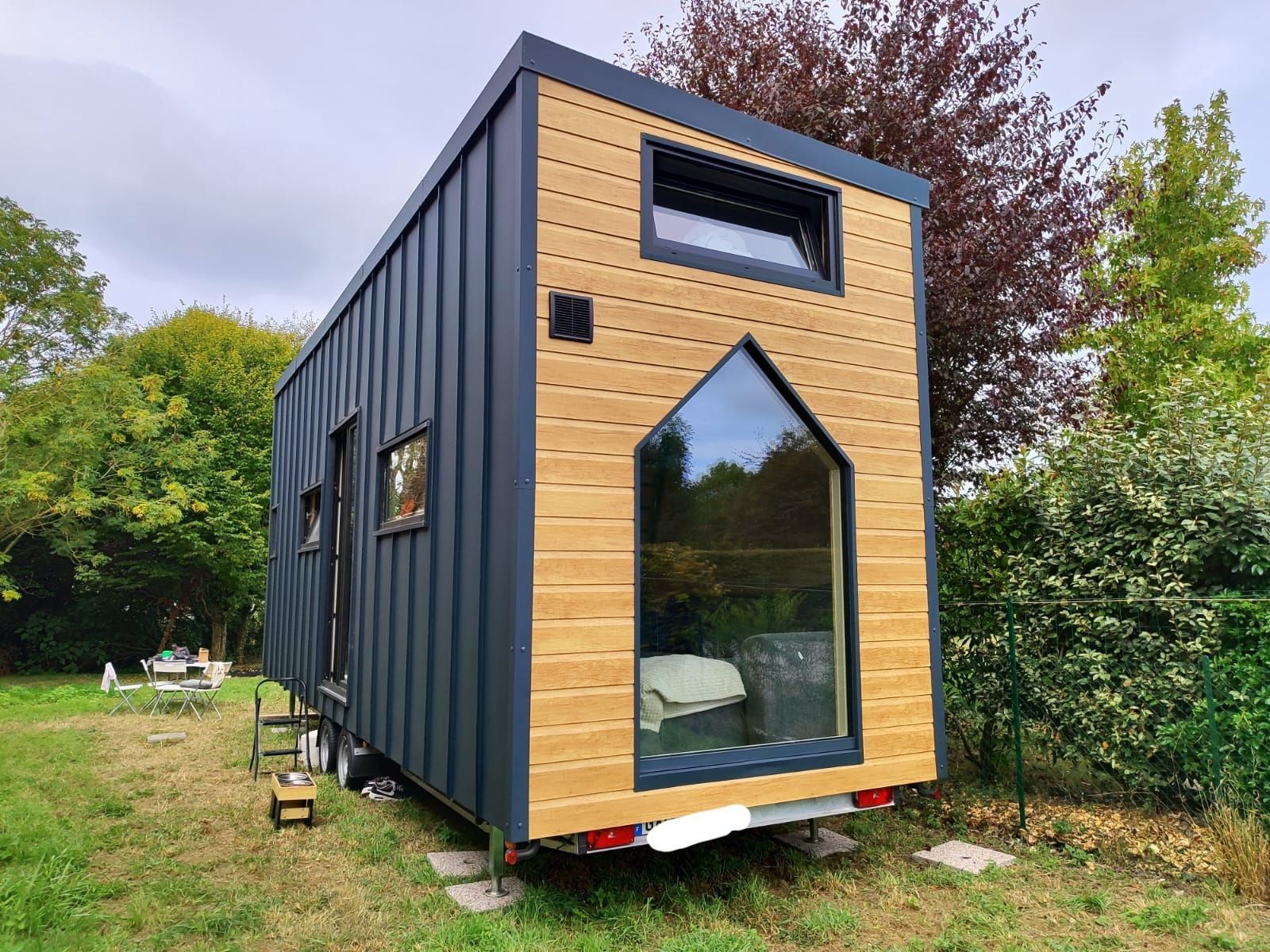 Petite maison moderne minuscule avec bardage noir et façade en bois, située sur un terrain herbeux à côté des arbres.
