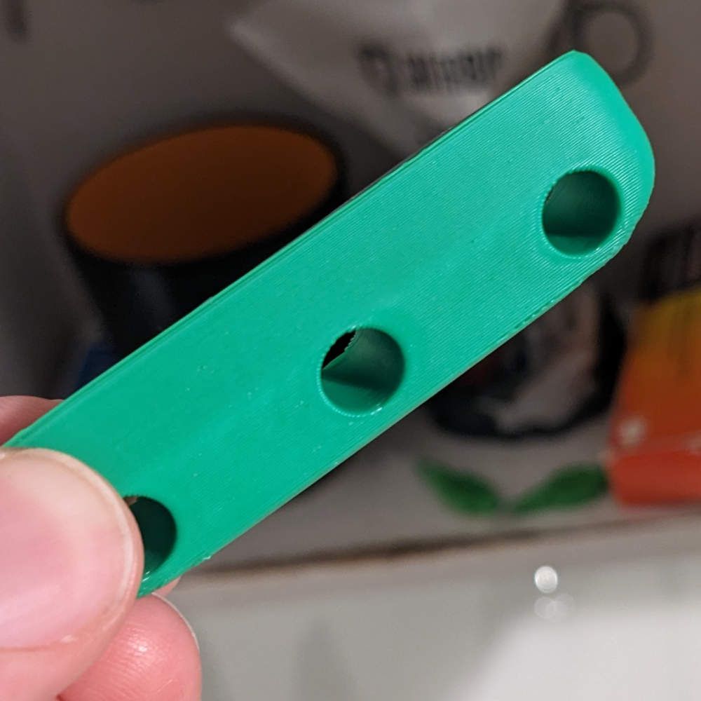 Grünes, 3D-gedrucktes Kunststoffteil mit drei Löchern, das von einer Hand gehalten wird.