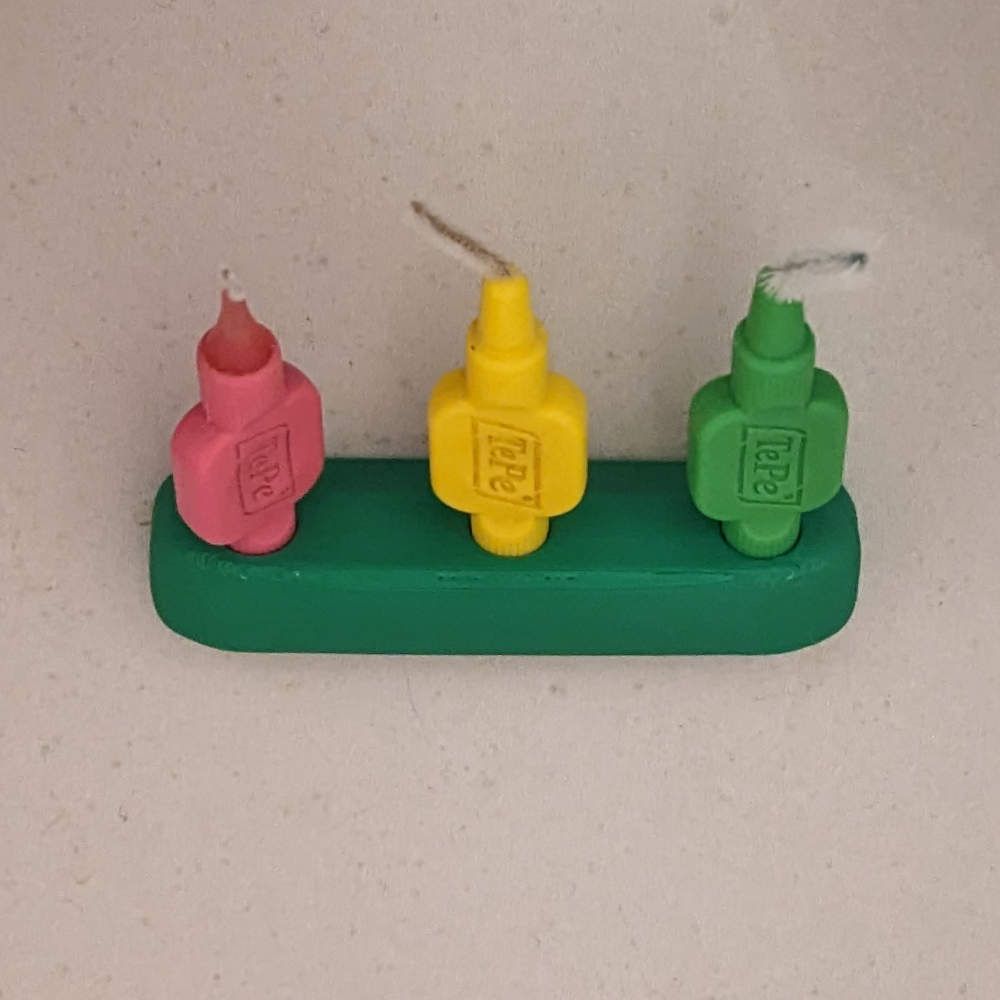 Drei Interdentalbürsten: rosa, gelb und grün, in einem grünen Halter auf einer gesprenkelten Oberfläche.