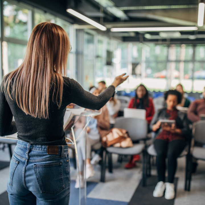 Mujer dando una presentación en un aula con estudiantes diversos.