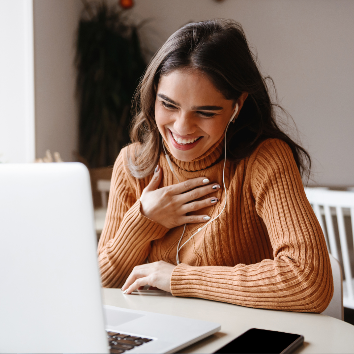 Una mujer con suéter naranja ríe mientras realiza una videollamada en una computadora portátil.