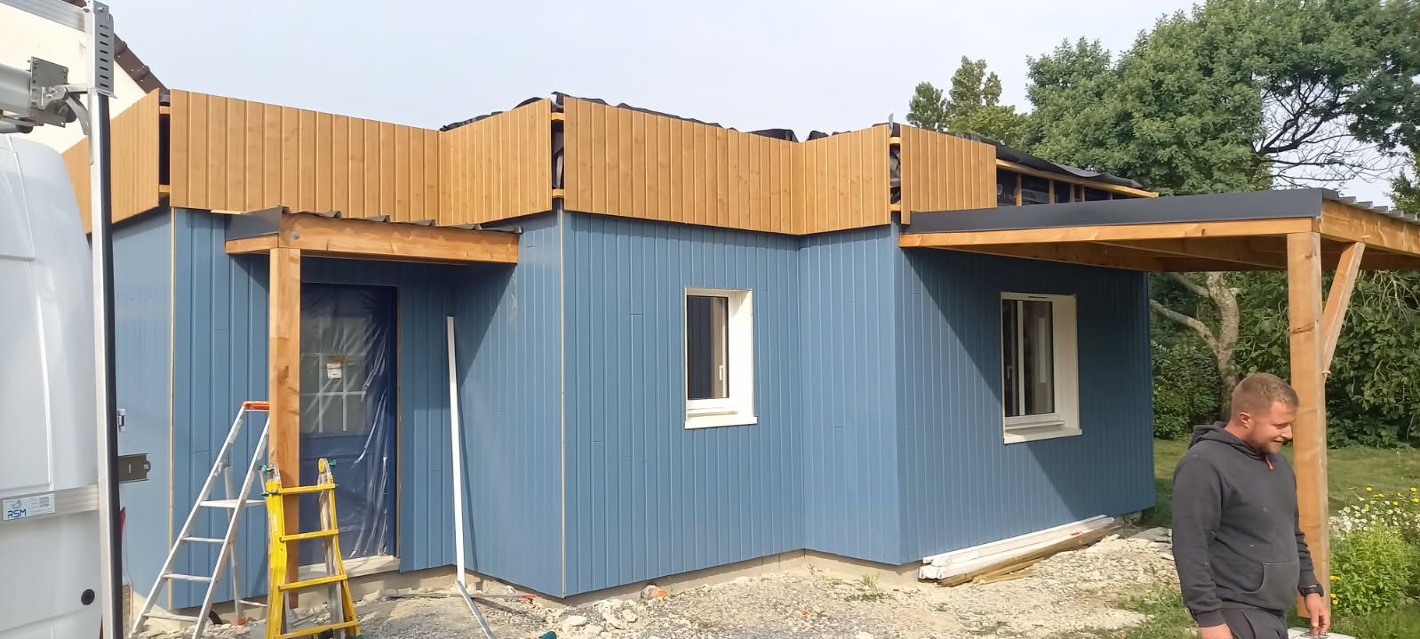 Cabane bleue aux accents de bois, un homme se tient près du porche, construction en cours.