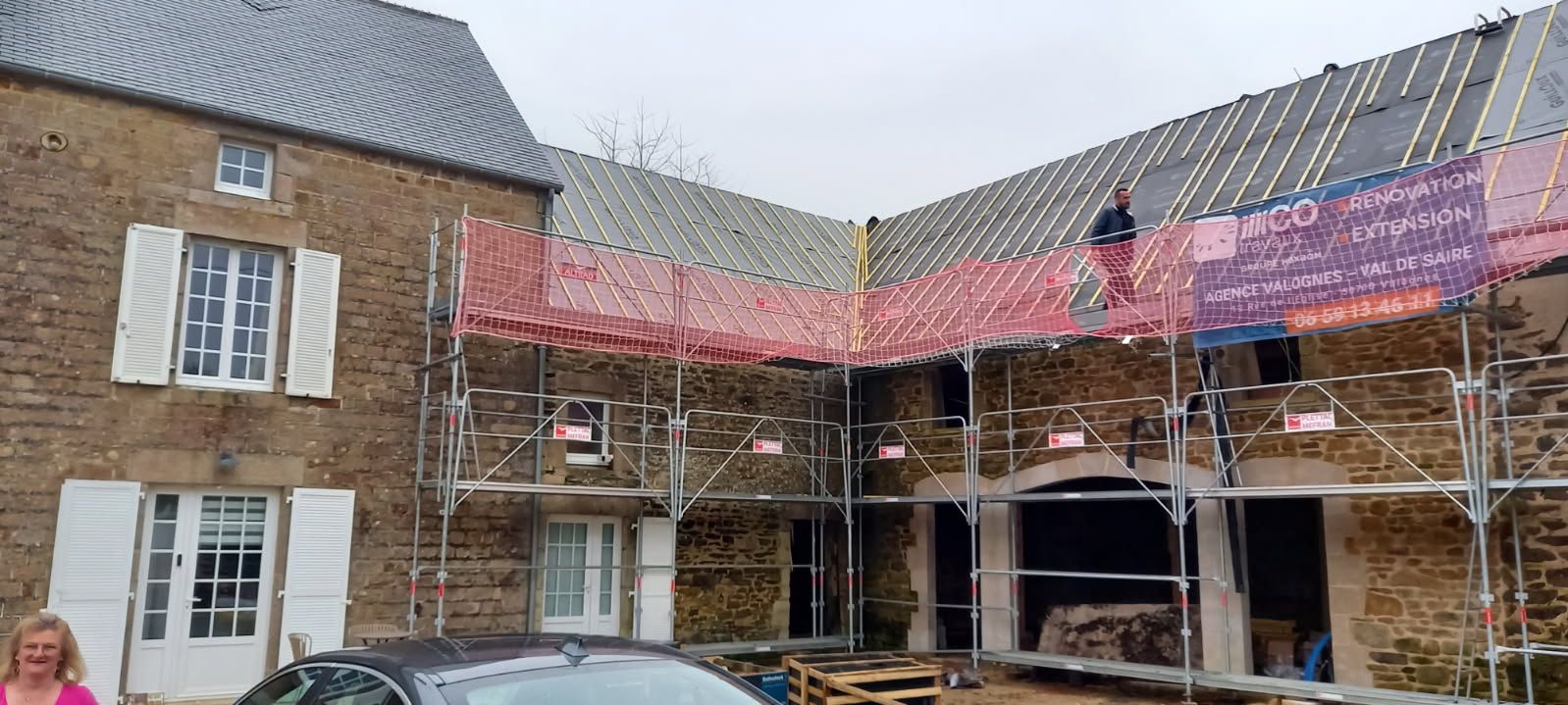 Extérieur du bâtiment avec échafaudage, toiture en cours de réparation. Façade en briques, filet de sécurité, personne sur le toit.