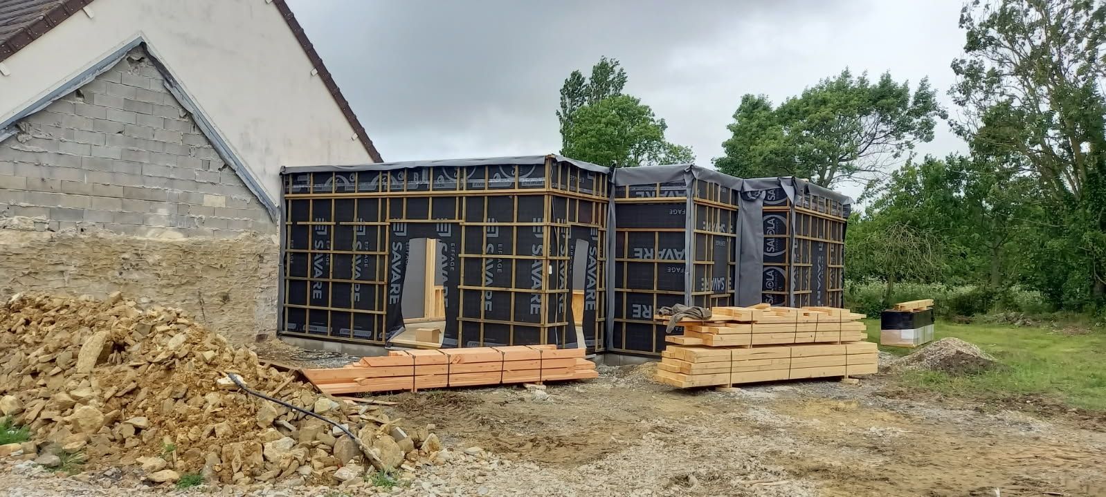 Chantier de construction avec ossature en bois pour nouvelle construction à côté d'une structure existante et de verdure.