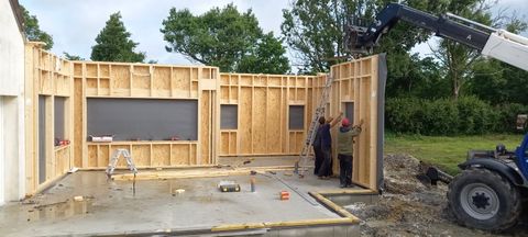 Des ouvriers du bâtiment installent une section de mur en bois sur une fondation en béton avec un chariot élévateur.