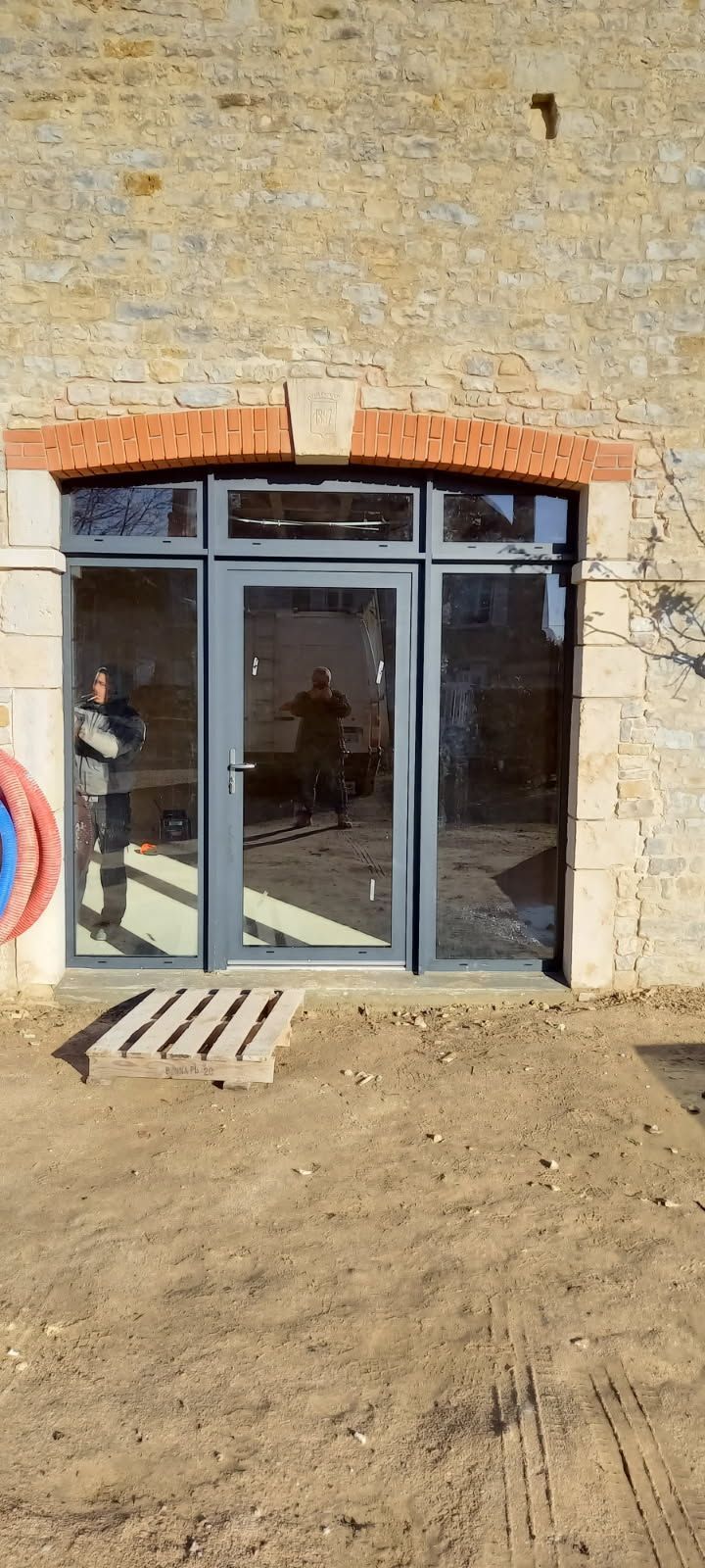 Porte vitrée dans un cadre gris, marches en briques, dessus en briques rouges, sur un mur en pierre beige.