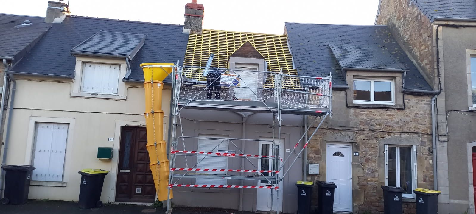 Maisons avec réparation de toiture en cours, échafaudages et chute à débris présents.