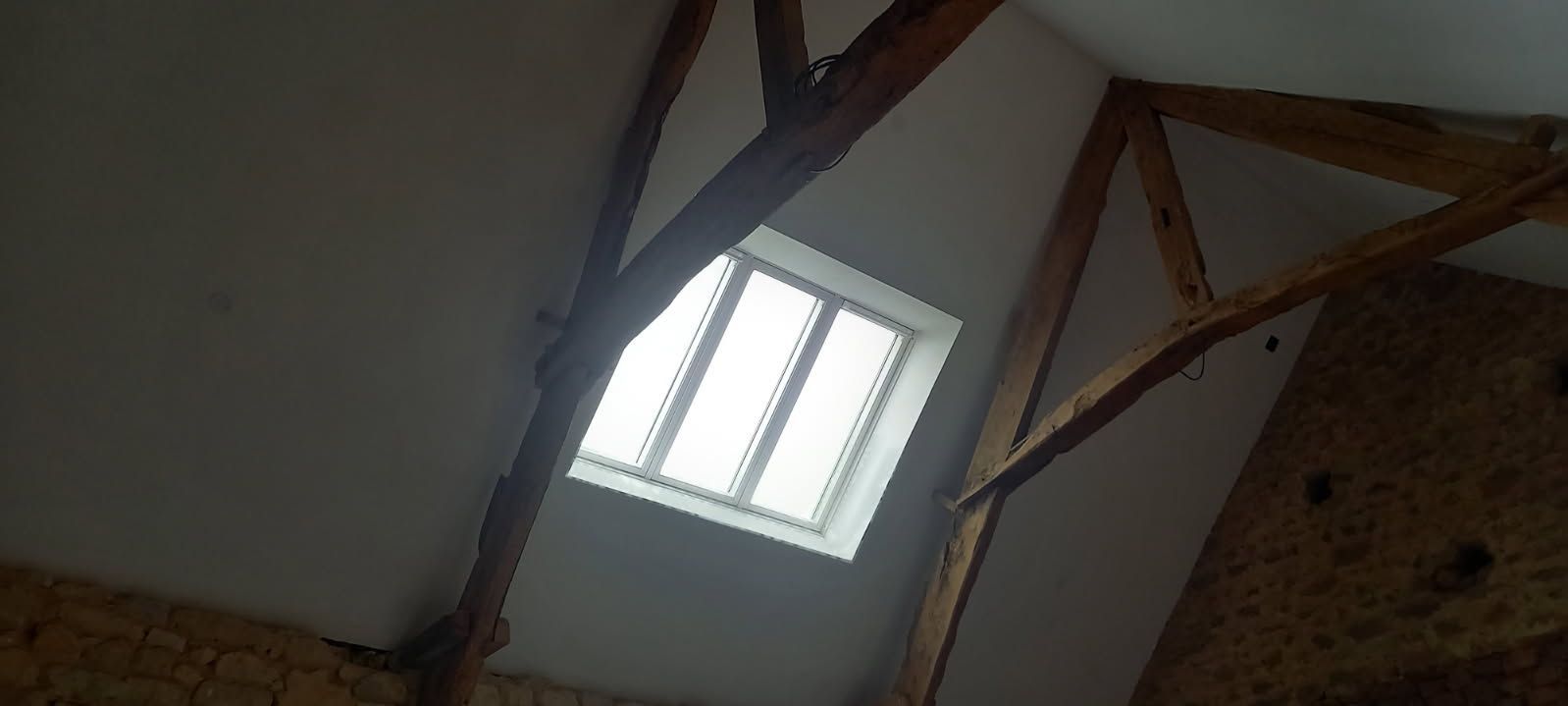 Vue intérieure d'une pièce avec poutres en bois et une fenêtre dans le haut plafond en angle.