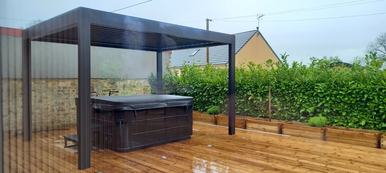 Jacuzzi sous une pergola marron. Terrasse en bois marron et haie verte en arrière-plan. Journée nuageuse.