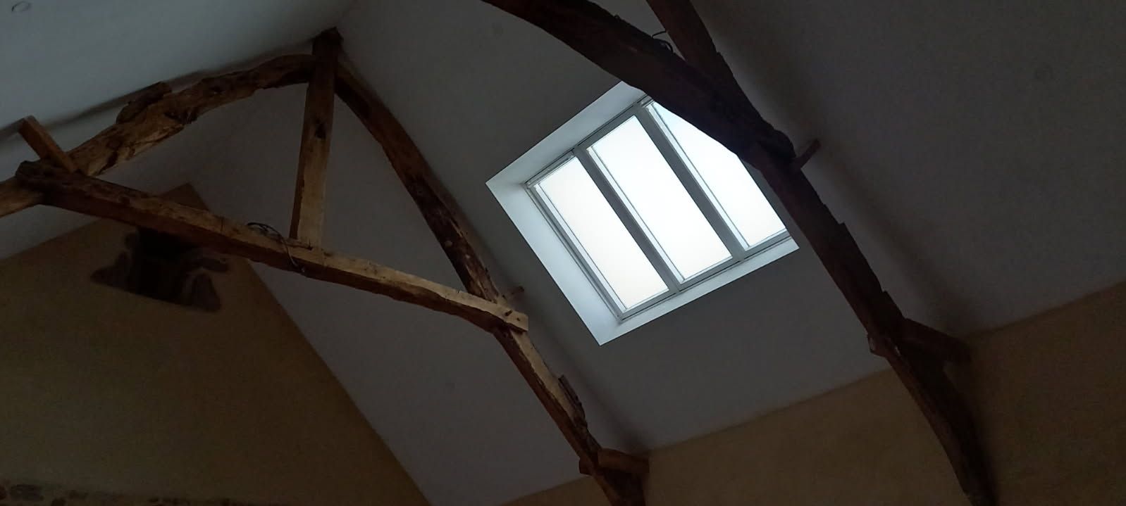 Vue intérieure d'un grenier avec puits de lumière et poutres en bois, les murs sont peints en beige.